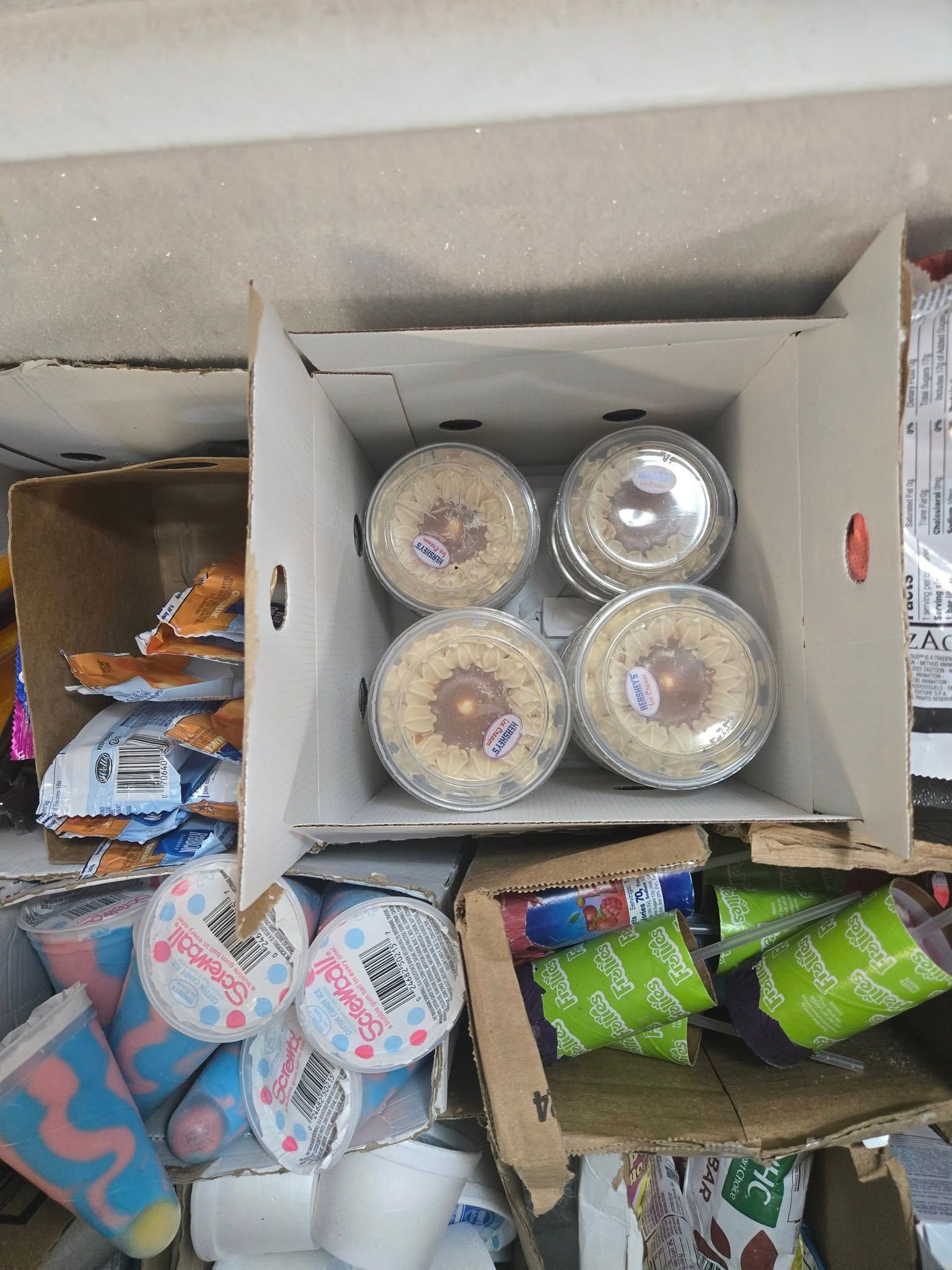 A cardboard box containing four small, circular, foil-topped desserts, surrounded by snack packaging in a retail setting.