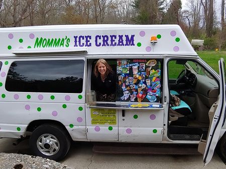 A smiling person leans out of the service window of a white ice cream van decorated with green and purple polka dots.