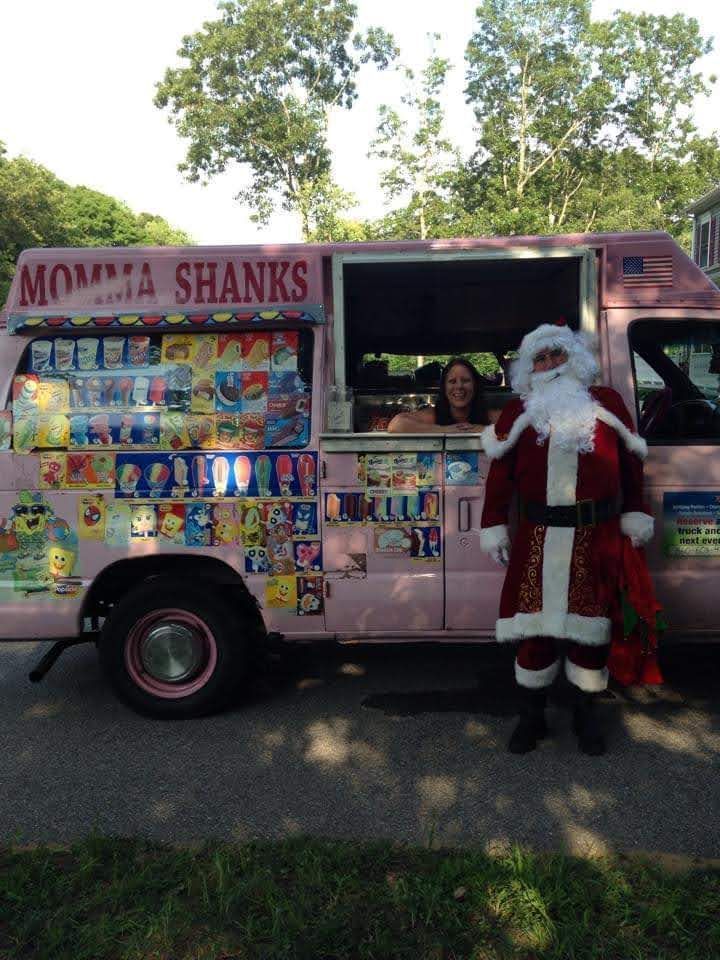 Santa Claus stands next to a pink Momma Shanks ice cream truck, while a smiling person looks out from the service window.
