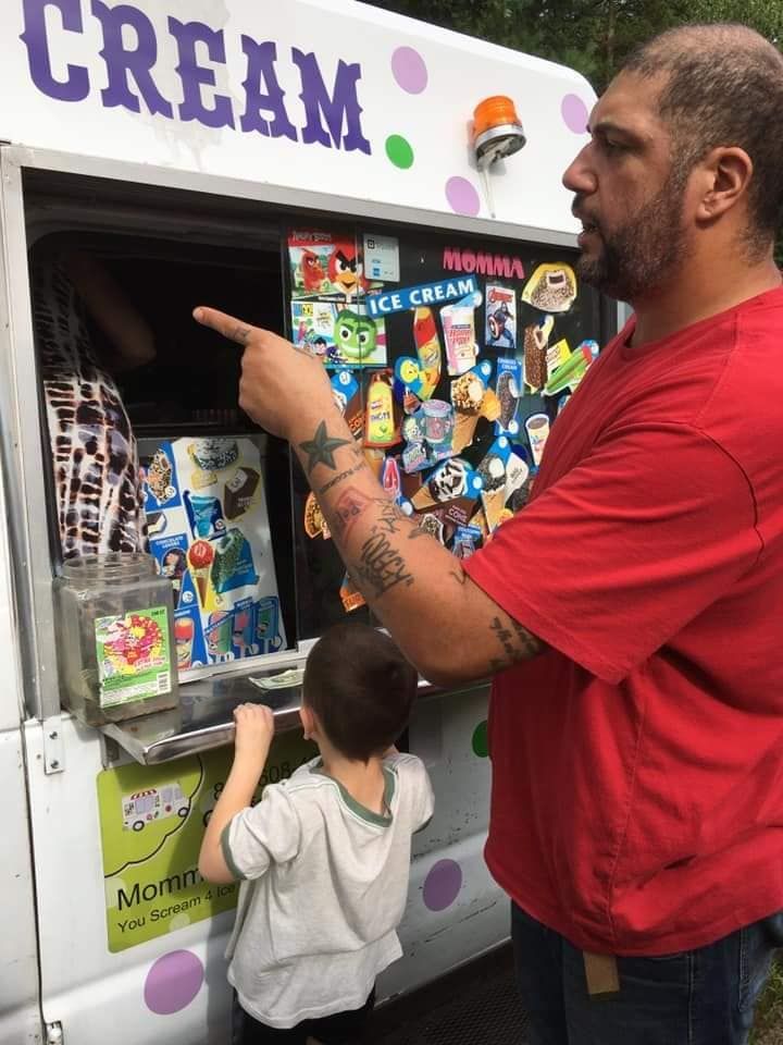 A person in a red shirt points to the menu of an ice cream truck while a child stands nearby looking at the selections.
