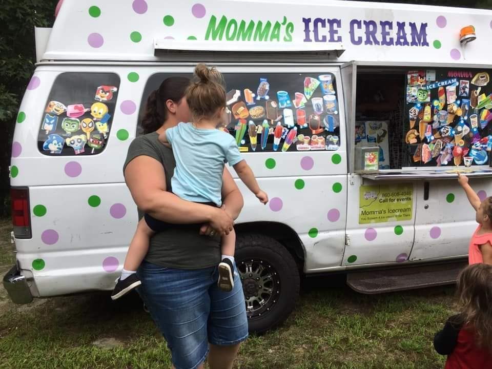A person holds a child outside a white ice cream truck with purple and green polka dots.
