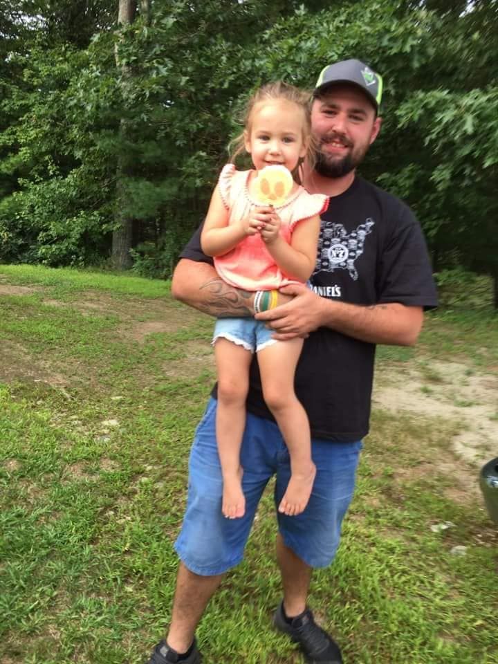 A person holds a child who is eating a lollipop outdoors in a wooded, grassy area.