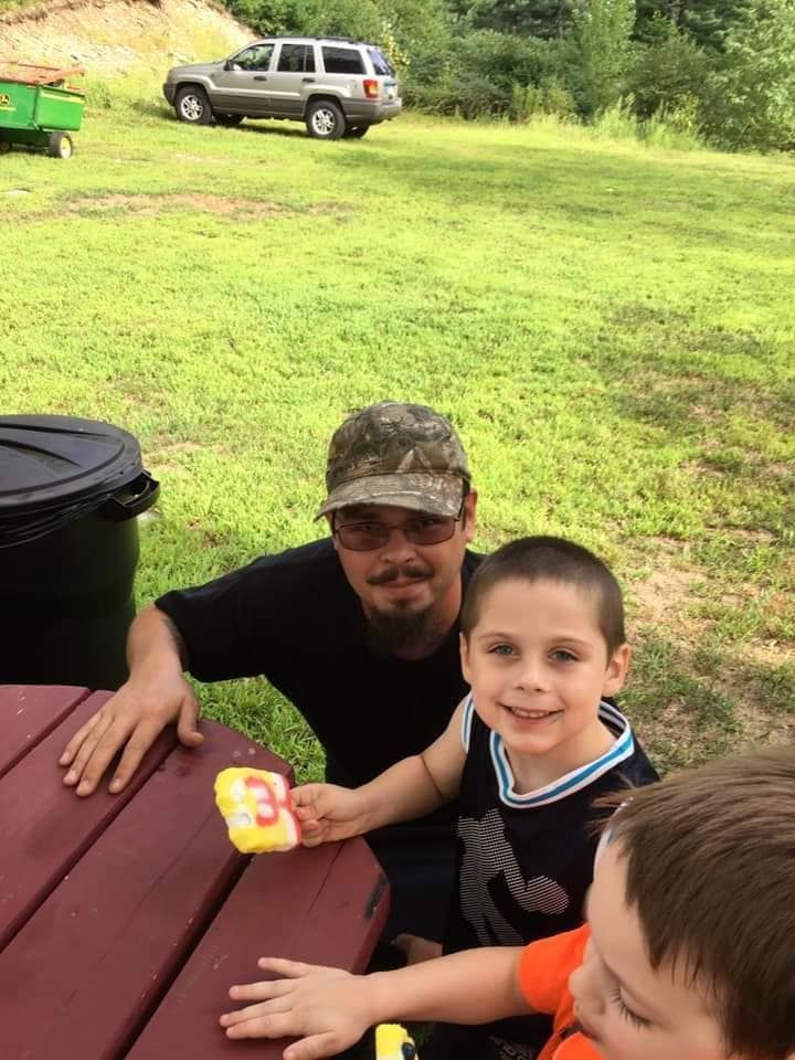 A person in a camouflage cap and two children eating popsicles at an outdoor table with a car parked on the grass behind.