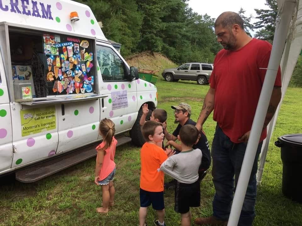 A group of children and an adult wait in line at a colorful ice cream truck parked on a grassy field.