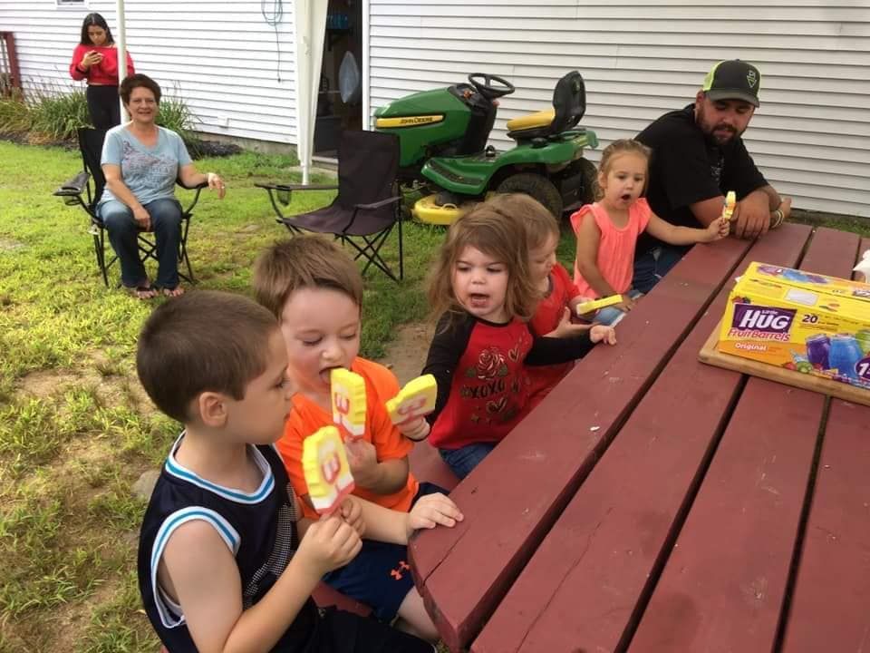 A group of children eating popsicles at a picnic table, with adults sitting nearby in a grassy, outdoor setting.