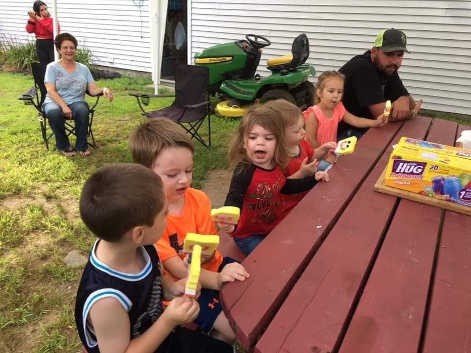 A group of children and adults sitting at an outdoor picnic table enjoying yellow popsicles on a sunny day.