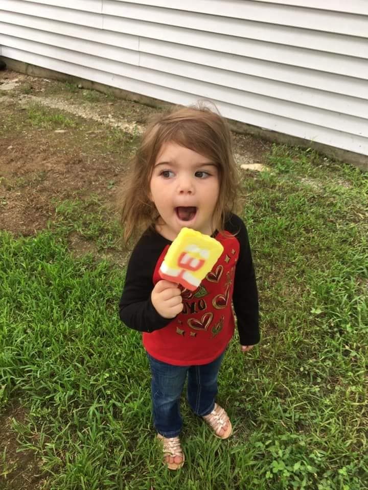 A young child standing on grass next to a house siding, eating a yellow and red popsicle with a surprised expression.
