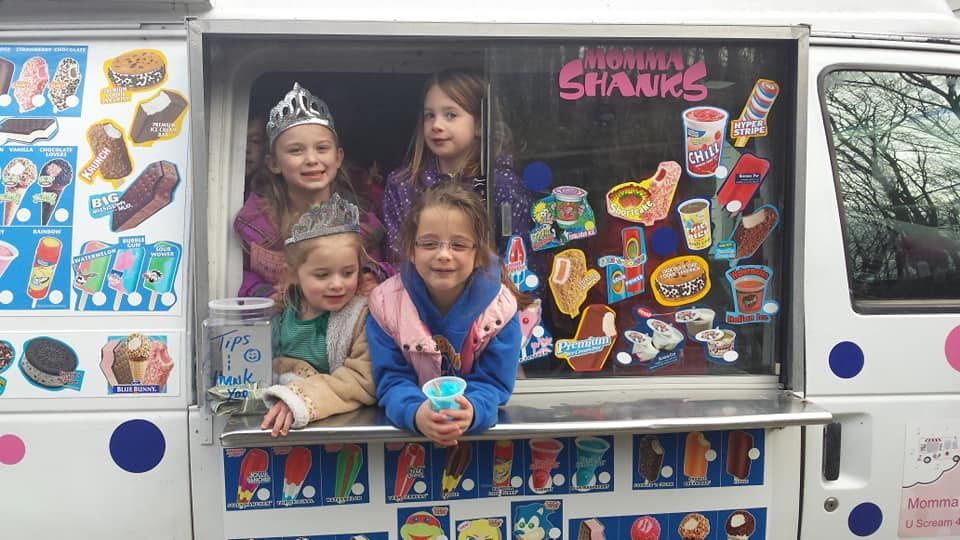 Four children smile while looking out from the service window of a white ice cream truck decorated with dessert graphics.