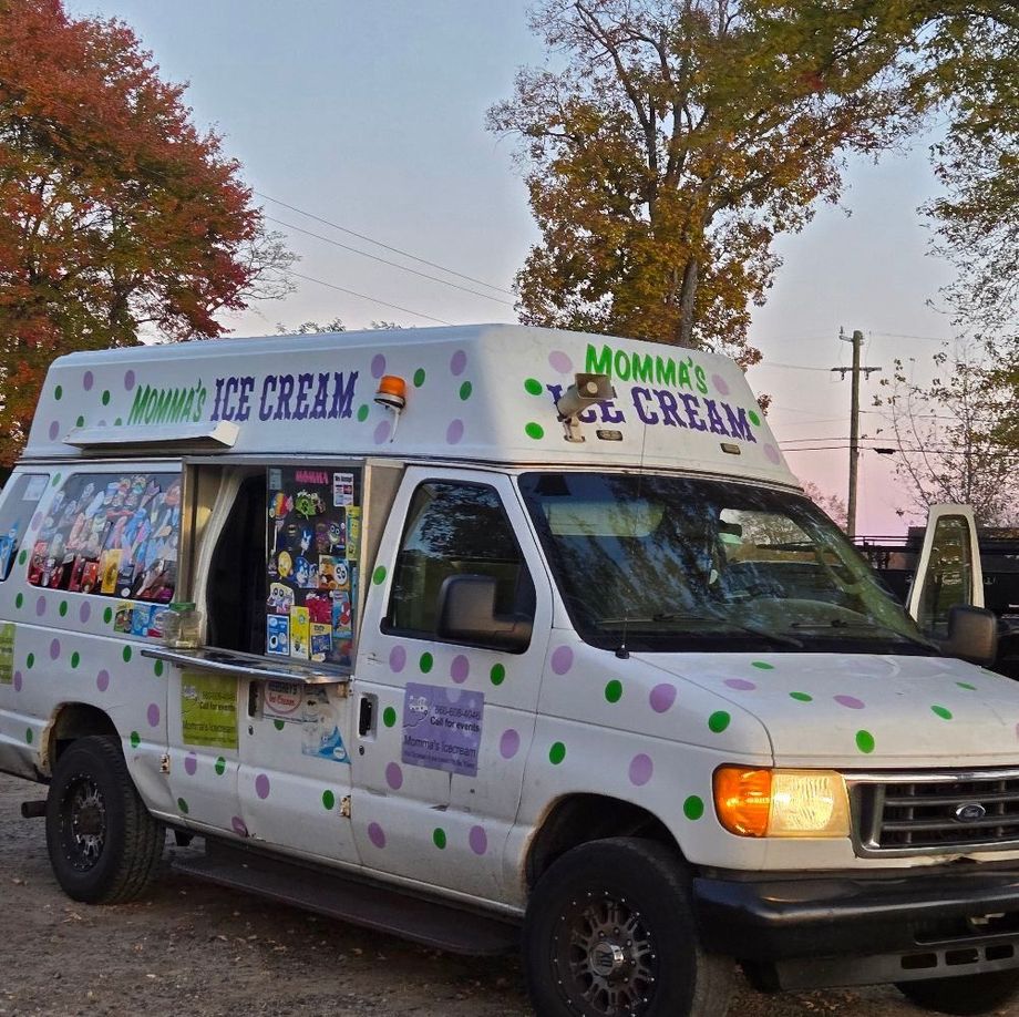 A white ice cream truck with purple and green polka dots parked outdoors under trees in the late afternoon.