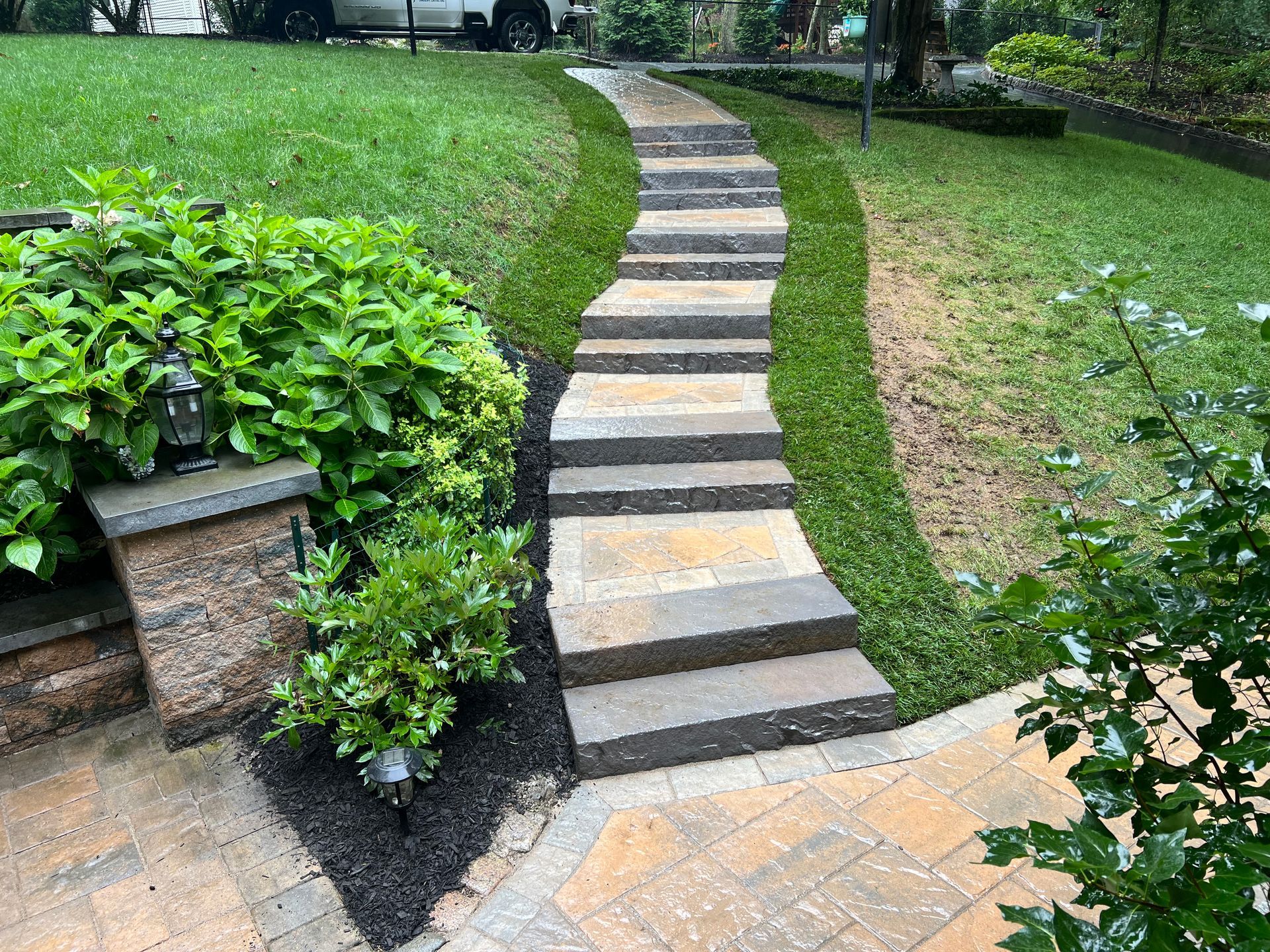 Stone steps ascend a sloped, grassy yard, flanked by dark mulch, greenery, and a paved walkway.