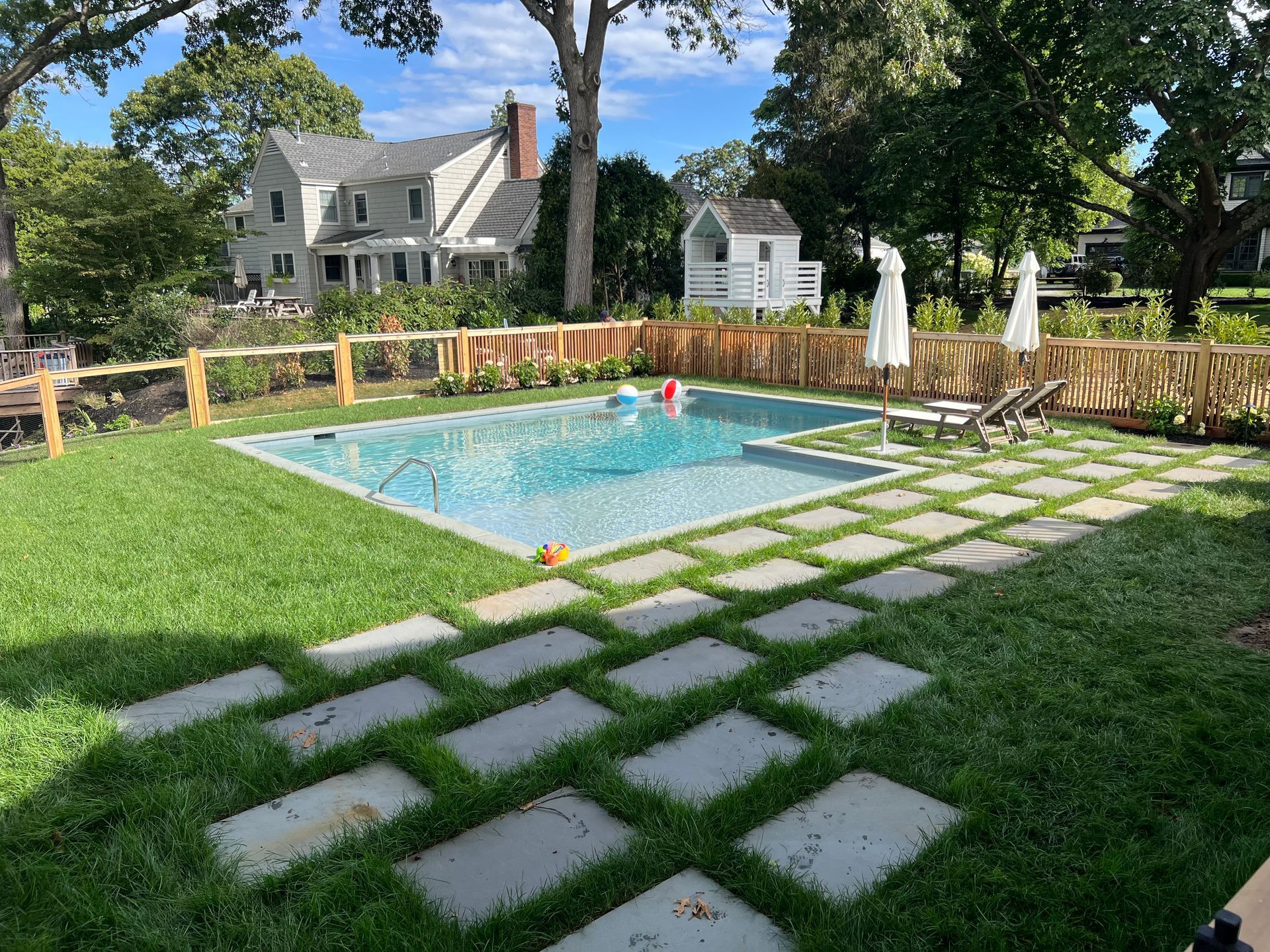 Backyard with a rectangular pool, stepping stone pathway, and a light-colored house in the background.