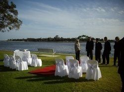 Wedding ceremony setup on a grassy waterfront, with white chairs, red carpet, and a few people standing.