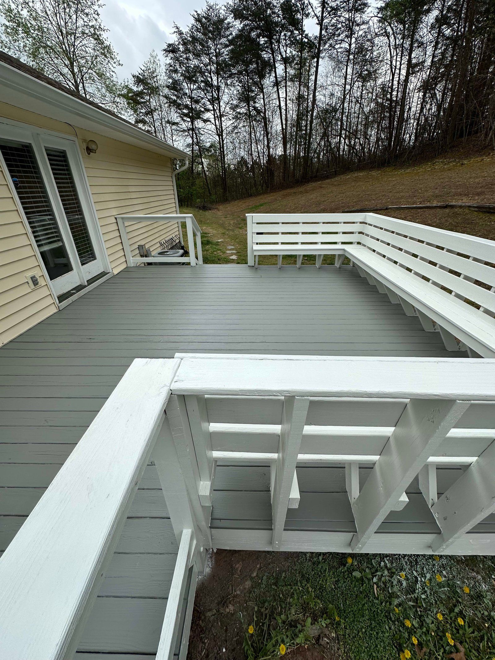 Gray-painted deck with white railing attached to a yellow house with a grassy hill and trees in the background.