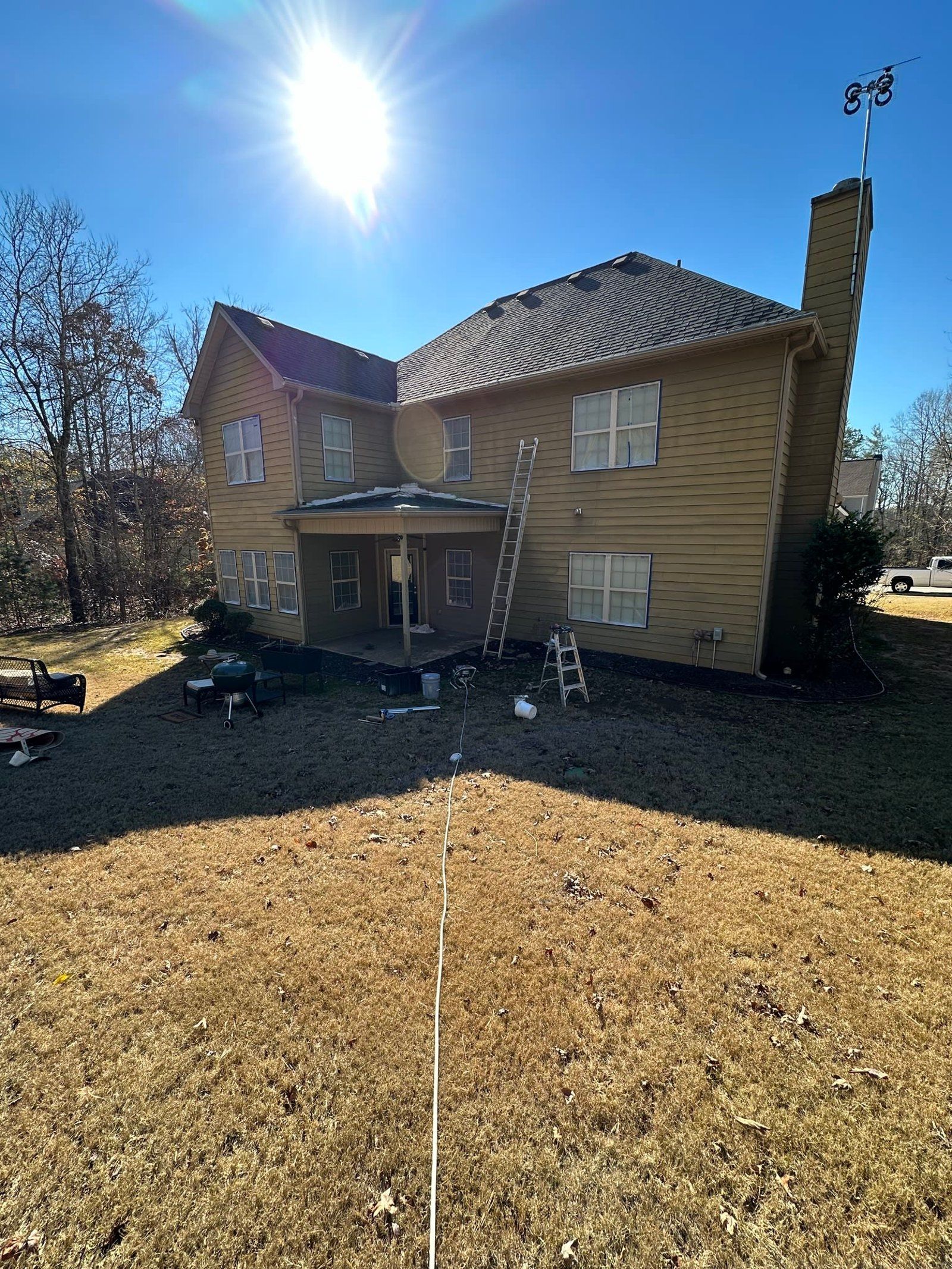 Two-story house with tan siding, chimney, and a ladder, under a bright sun.