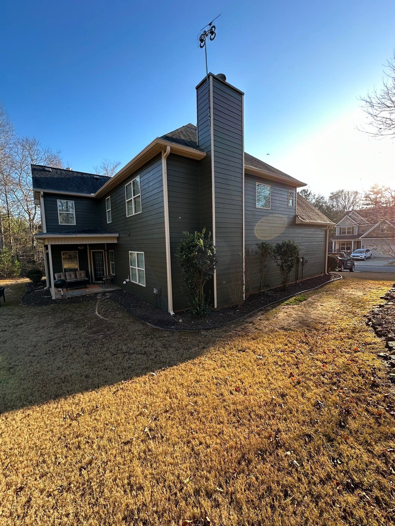 Dark green house with chimney under a sunny blue sky. Brown grass in the yard.