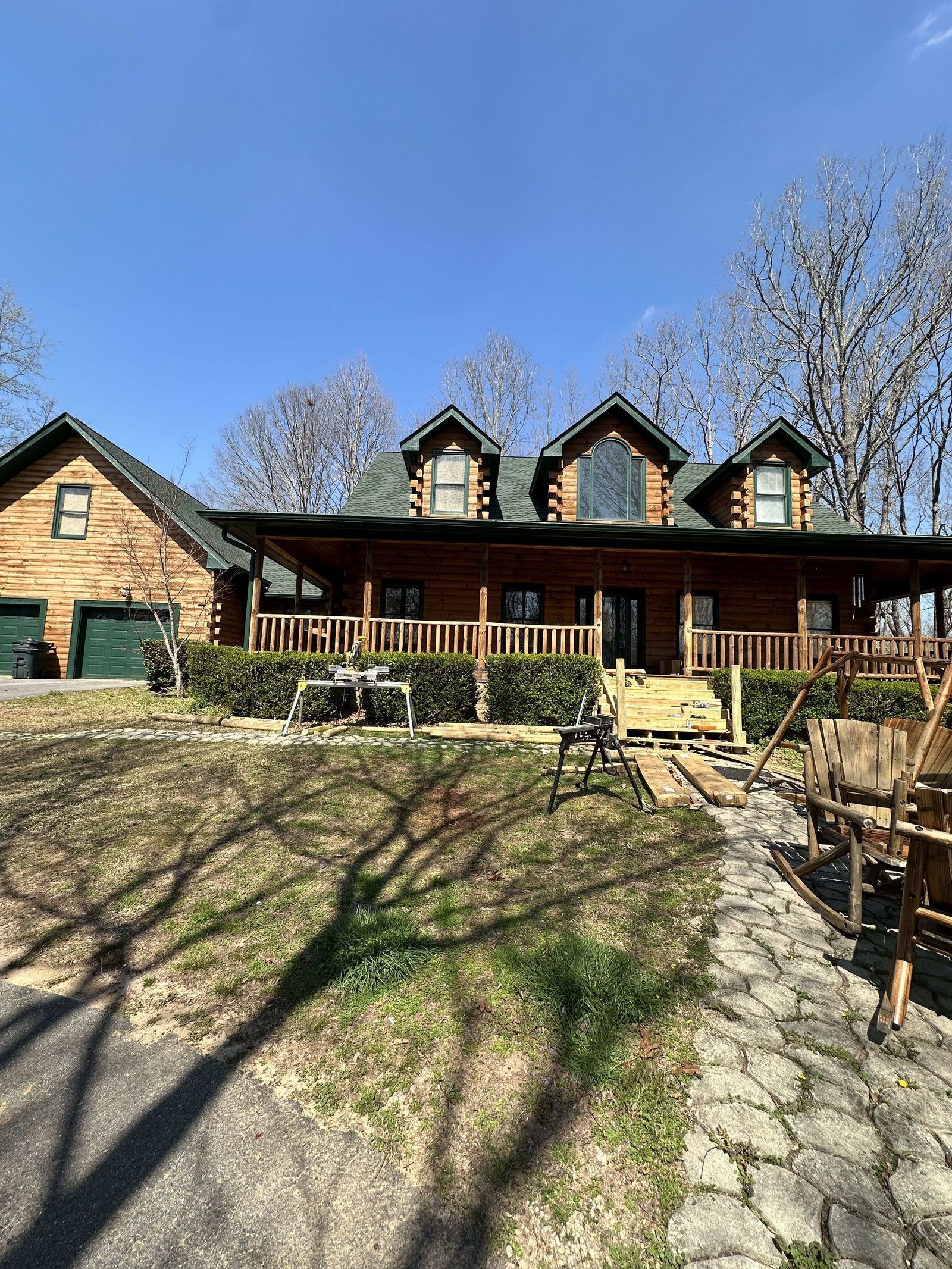 Log cabin house with covered porch, landscaping, and detached garage on a sunny day.