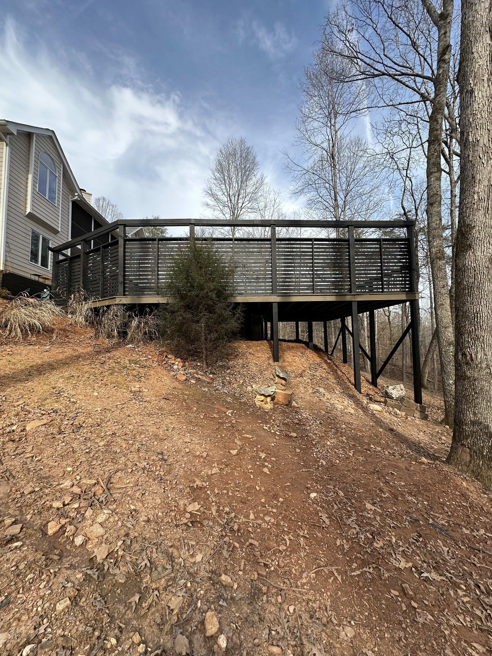 Elevated wooden deck painted black on a sloped, dirt yard with surrounding trees and a house.