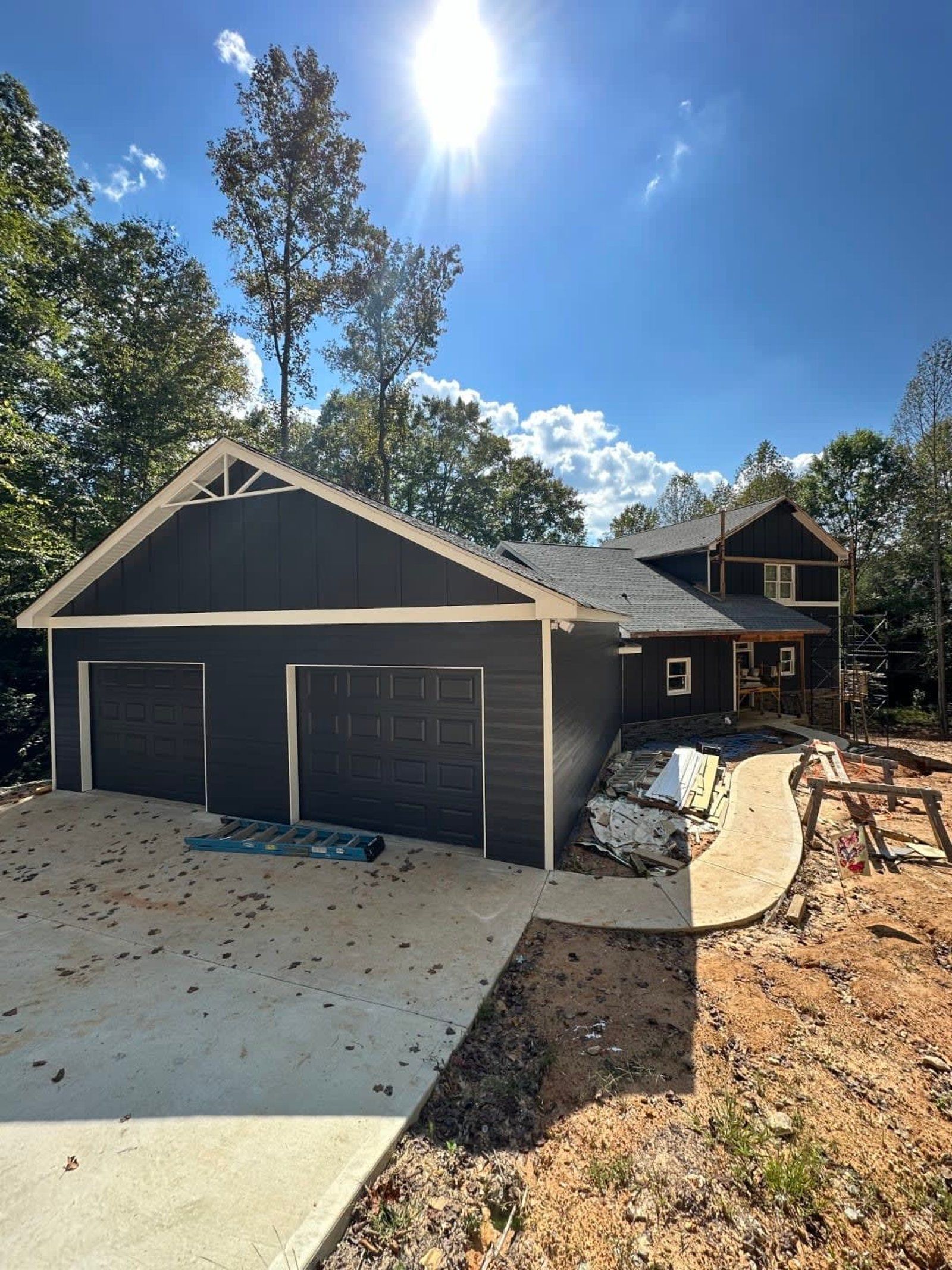 Dark gray garage with three doors, bright sky, concrete driveway, and construction materials.