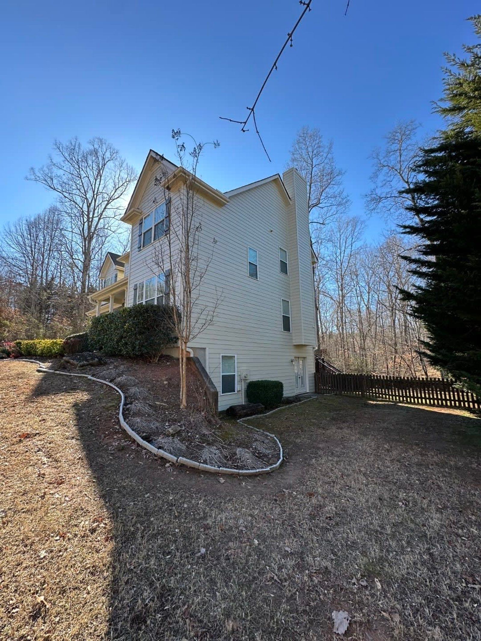 Beige two-story house on a hillside, surrounded by trees and a brown yard under a clear blue sky.