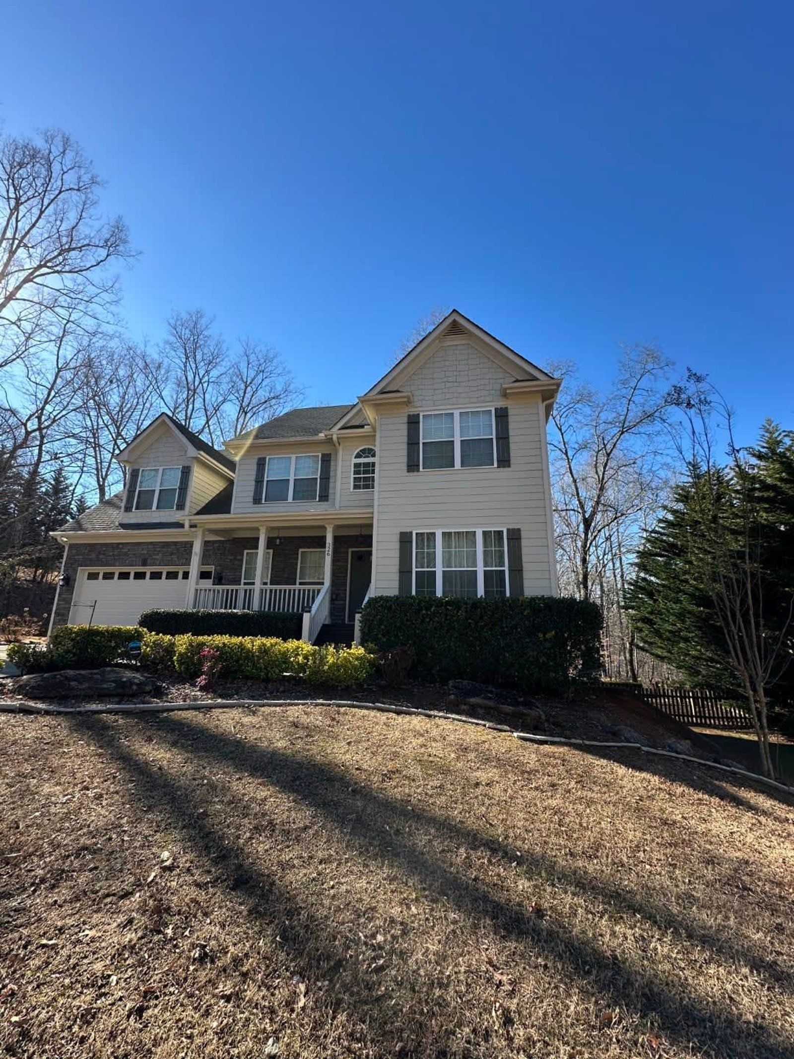 Two-story beige house with green shutters on a sunny day. Brown grass and trees in the foreground.