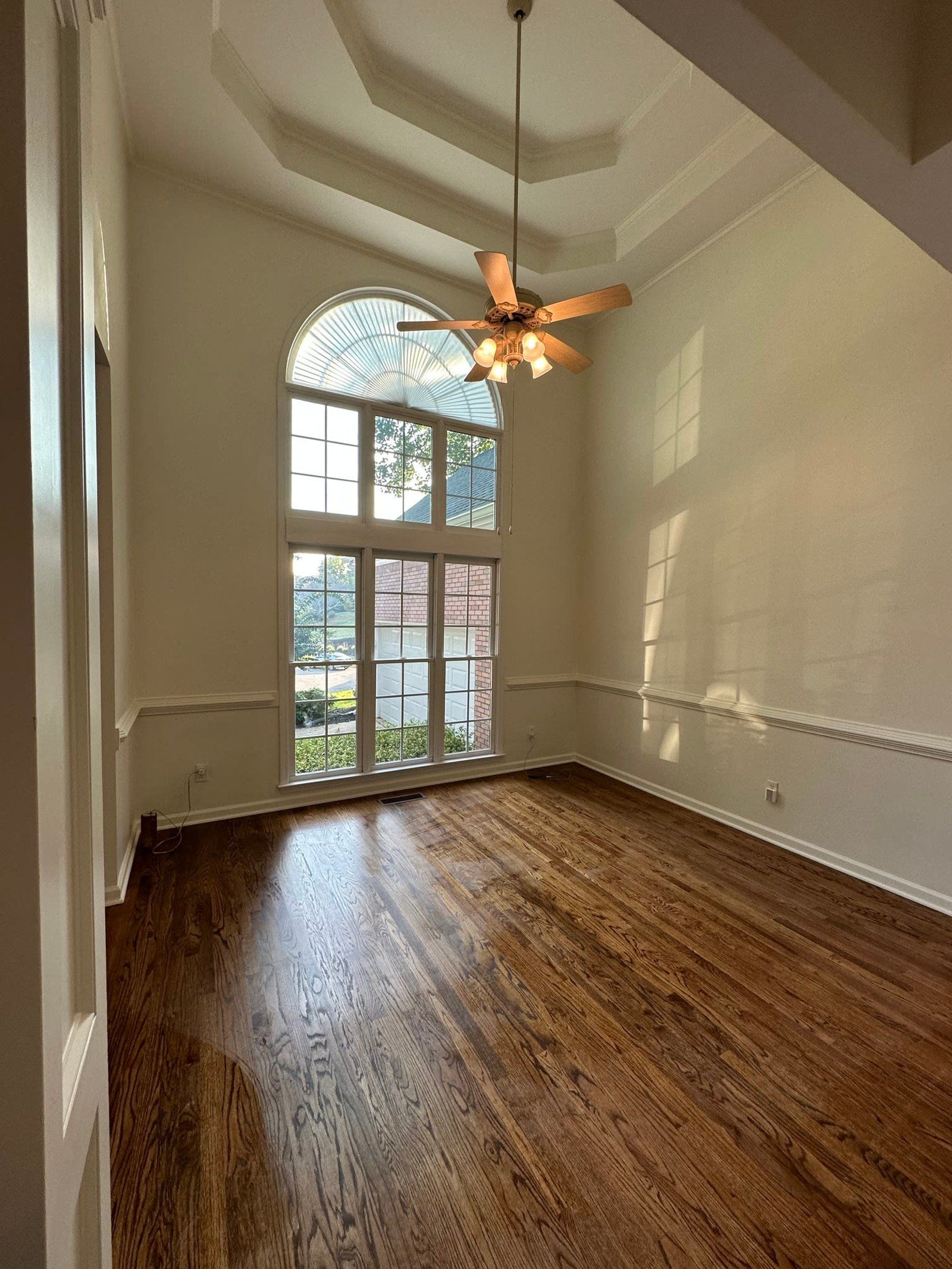 Empty room with hardwood floors, tall arched window, and ceiling fan.