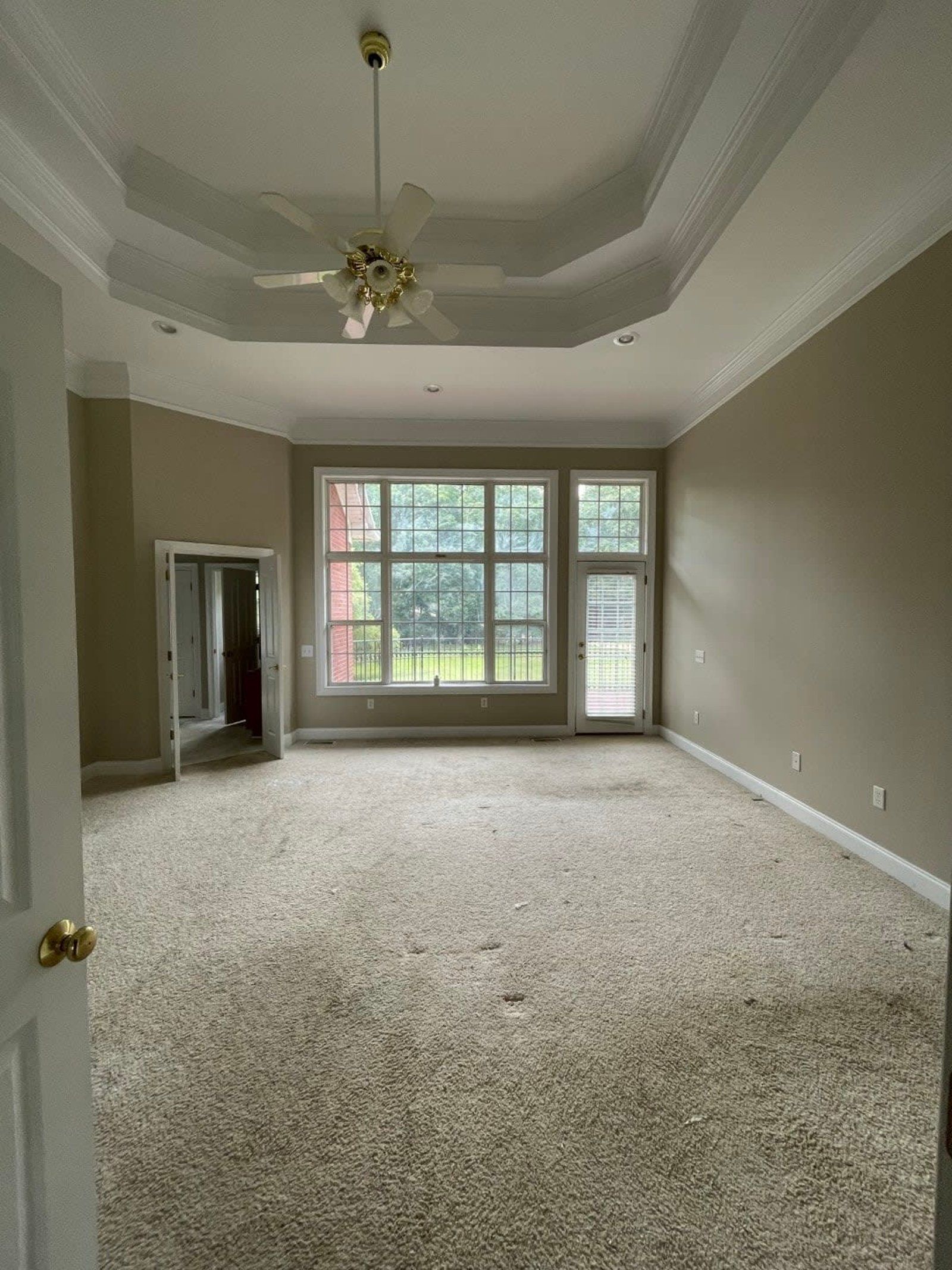 Empty bedroom with beige walls, carpet, and large window. Ceiling fan and tray ceiling.
