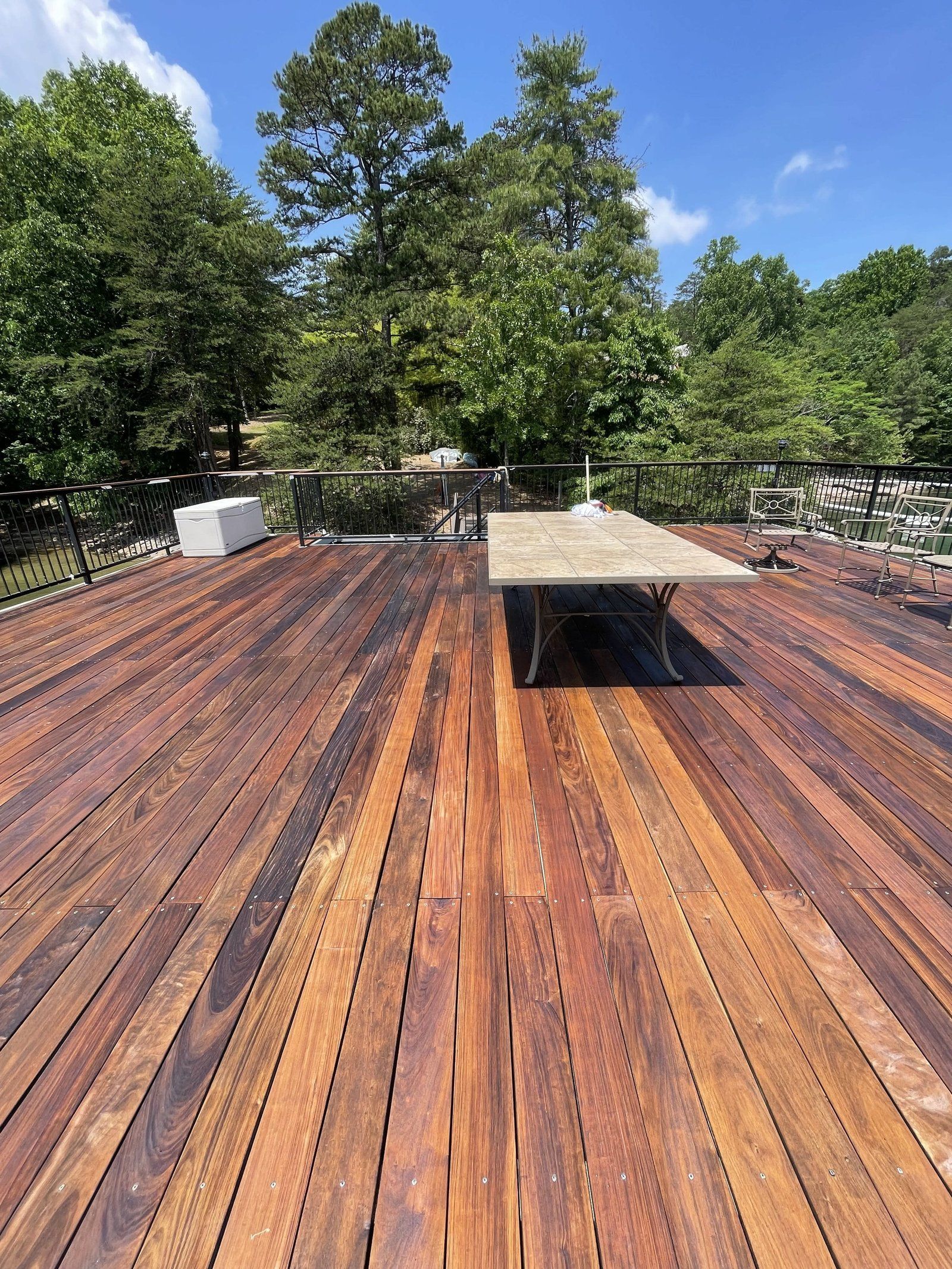 Wooden deck with a table, surrounded by trees under a blue sky.