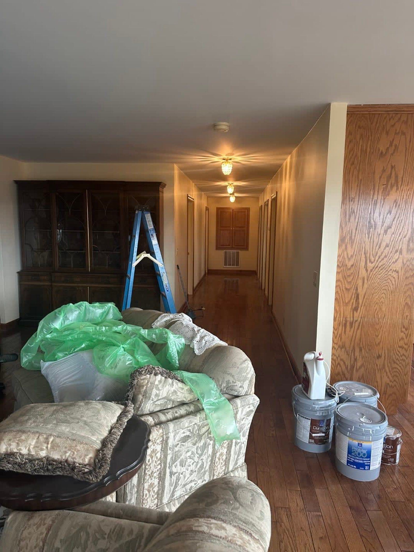 Renovated hallway with a ladder, paint buckets, and covered furniture; wooden floors.