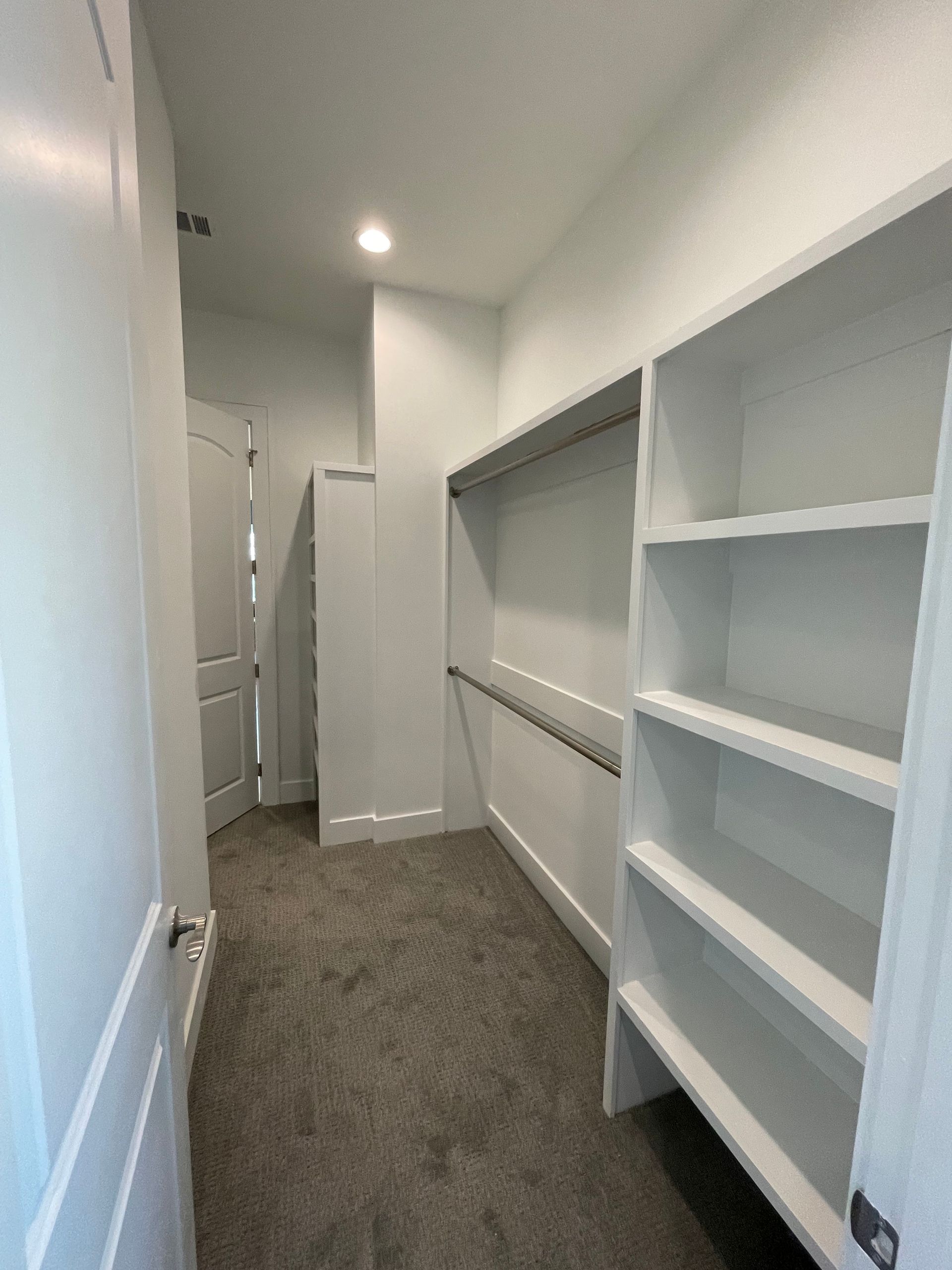 Empty walk-in closet with white shelves, hanging rod, door, and gray carpet.