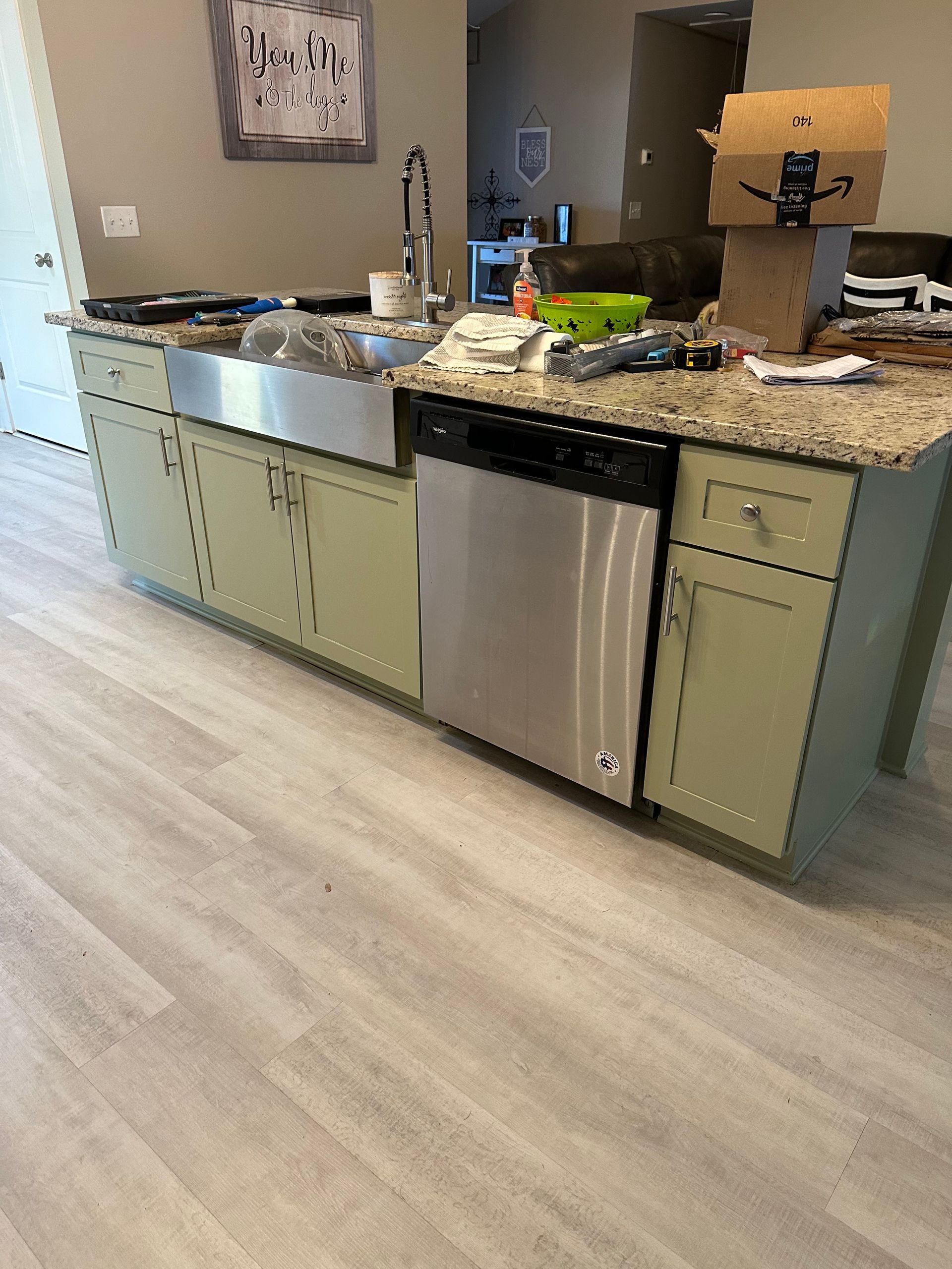 Kitchen island with stainless steel sink and dishwasher, light green cabinets, granite countertop, and light wood-look floor.