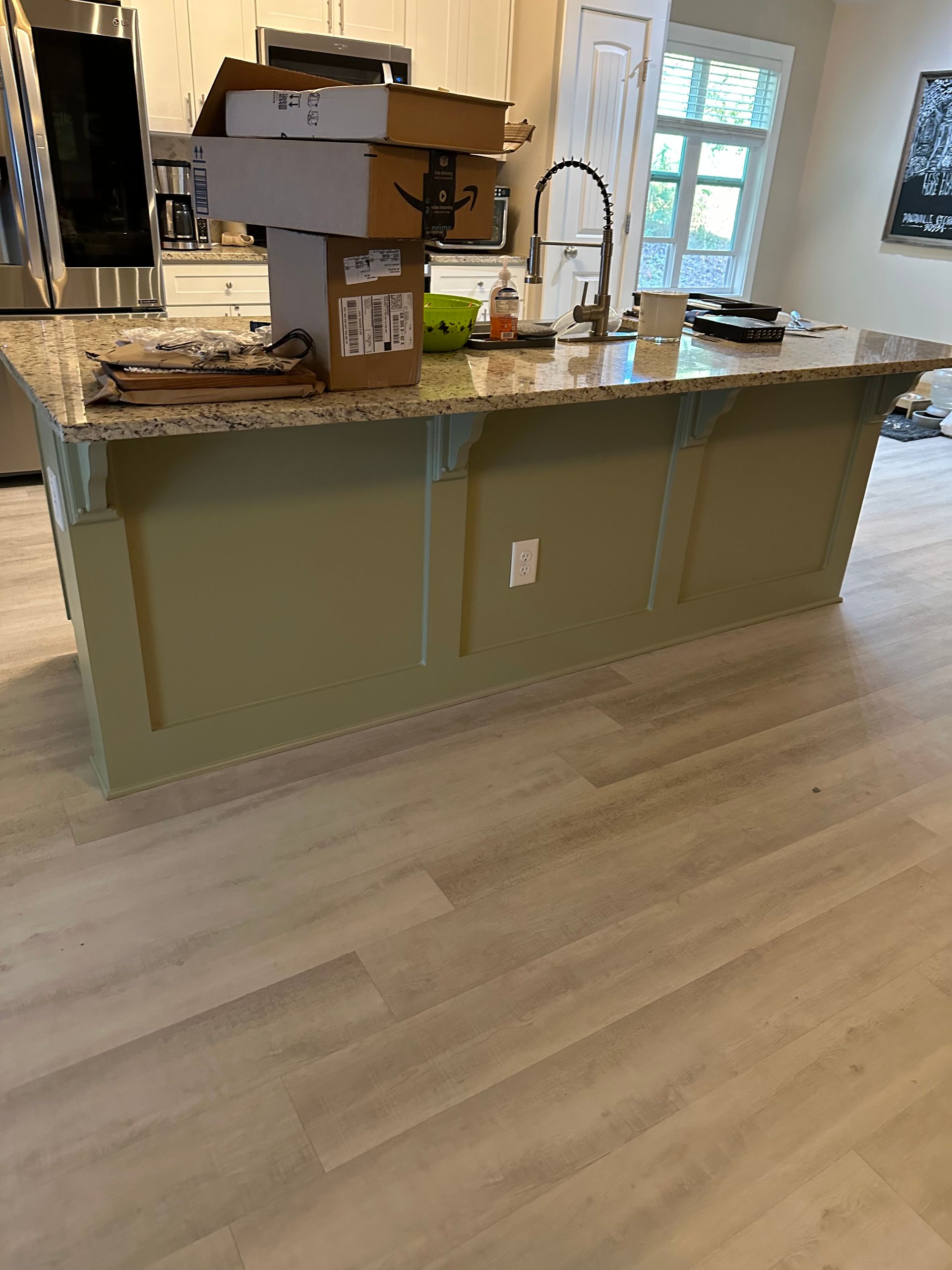 Kitchen island with granite countertop, painted green base, and light wood-look flooring. Amazon boxes on top.
