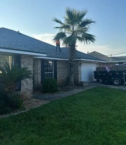 Person on a roof, near a palm tree, working on shingles. Brick house with a trailer in driveway.