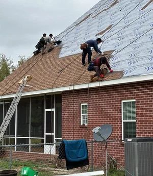 Roofers installing shingles on a brick house with a cloudy sky backdrop.