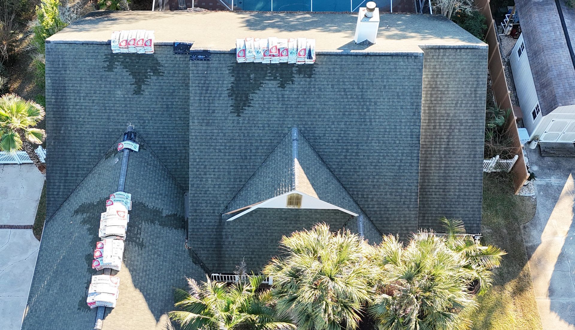 Overhead view of a house roof with dark shingles. Sections covered with tarps; trees below.