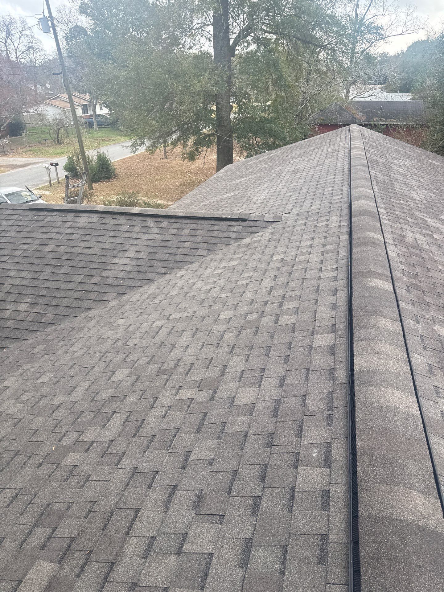 A high-angle view of a gray shingled roof with a central ridge, extending out to a residential street and trees.