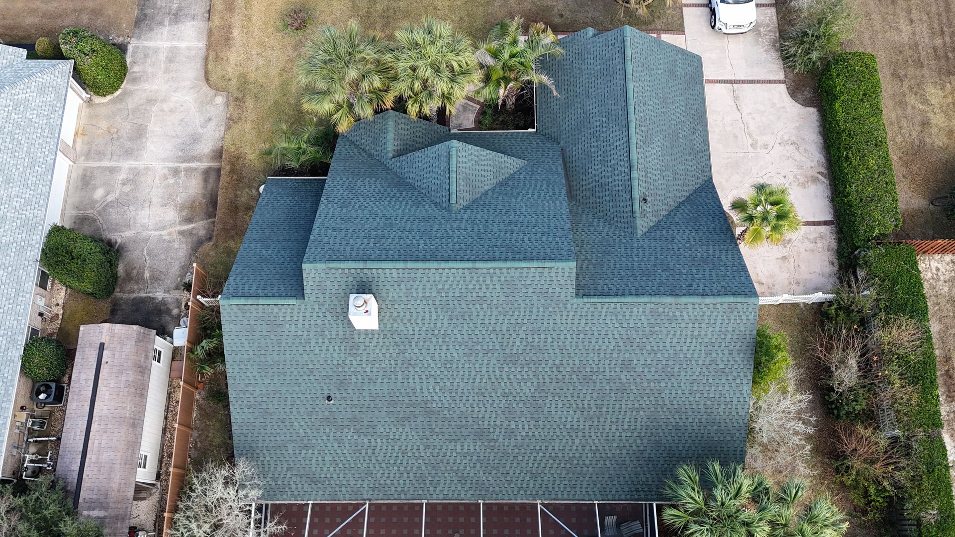 An aerial view of a green shingled residential roof with multiple peaks, a chimney, and adjacent landscaped driveways.
