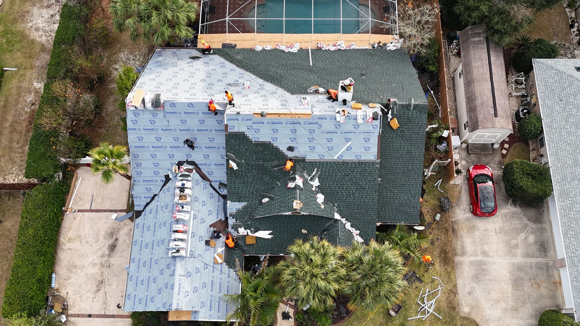 Roofers repairing a house roof. Aerial view shows workers, materials, and a swimming pool in the background.