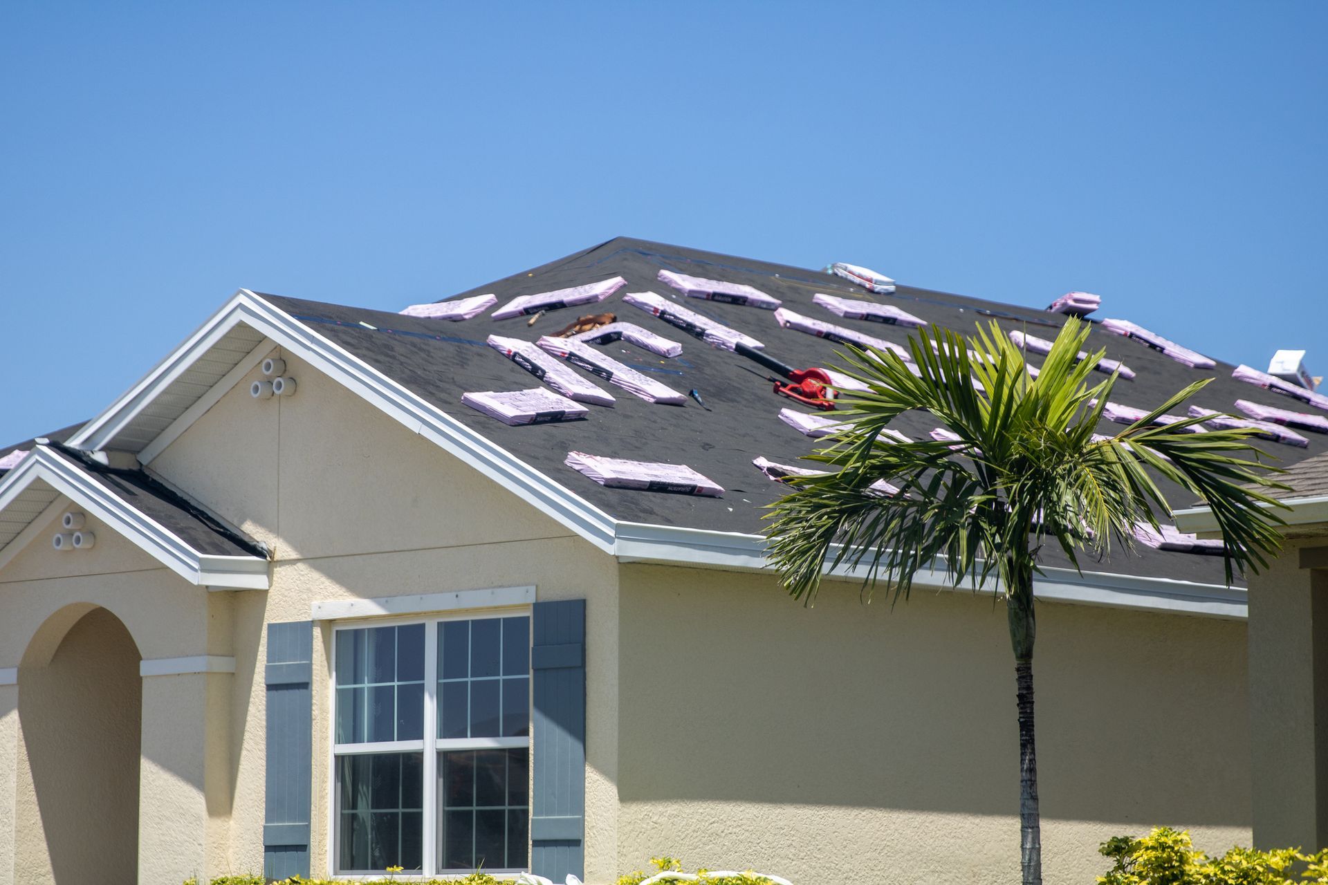 A house with roof shingles missing; pink insulation visible. Palm tree in front. Bright blue sky.