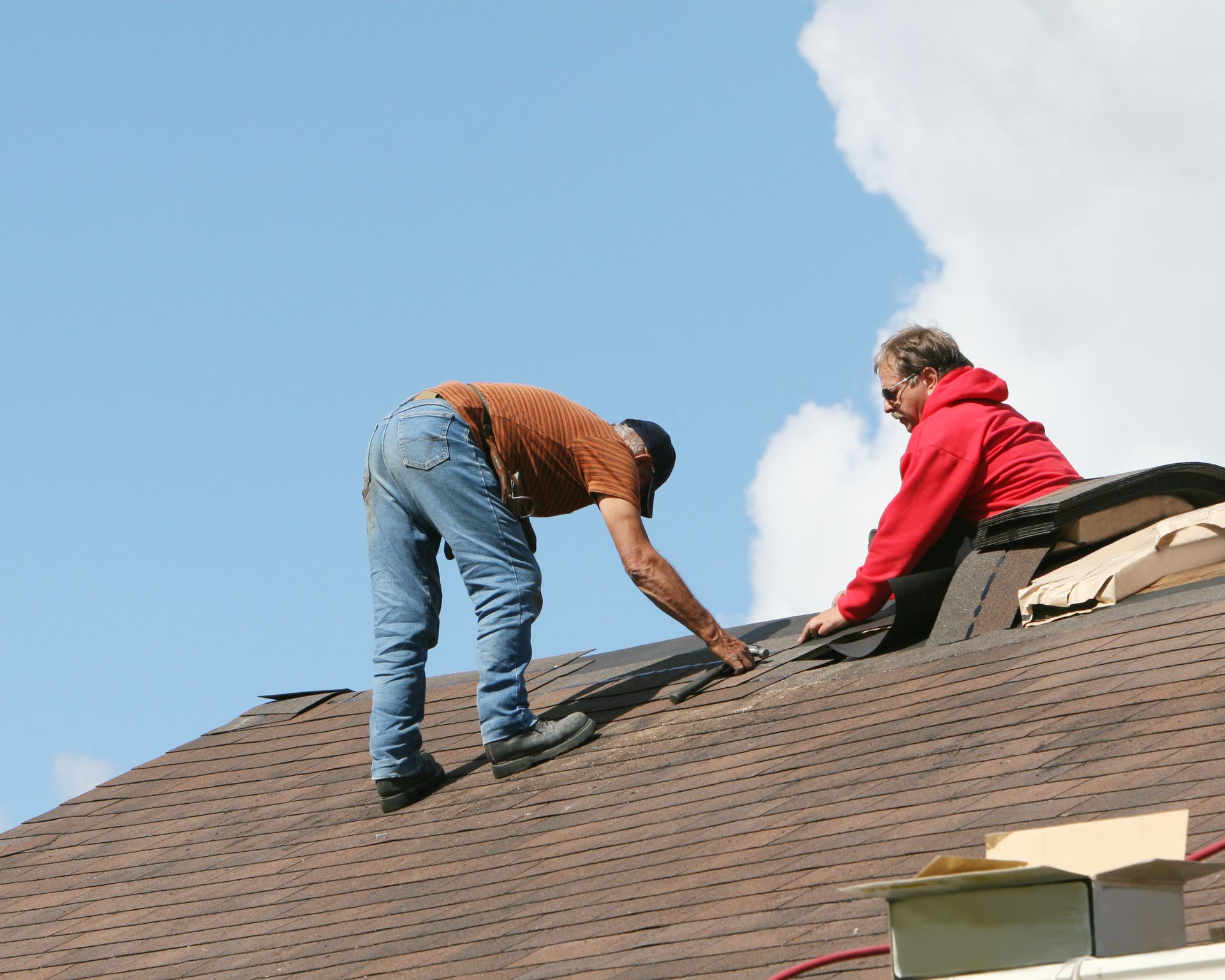 Two people on a brown shingled roof, one reaching, the other handling materials, against a blue sky with clouds.