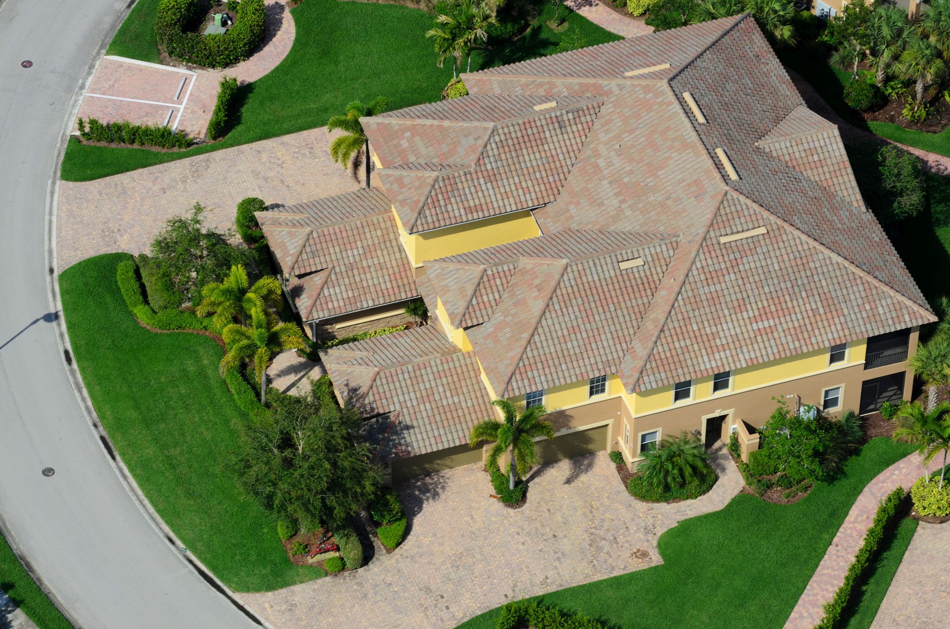 Aerial view of a large house with a terracotta tile roof, yellow walls, and a circular driveway on a well-manicured lawn.