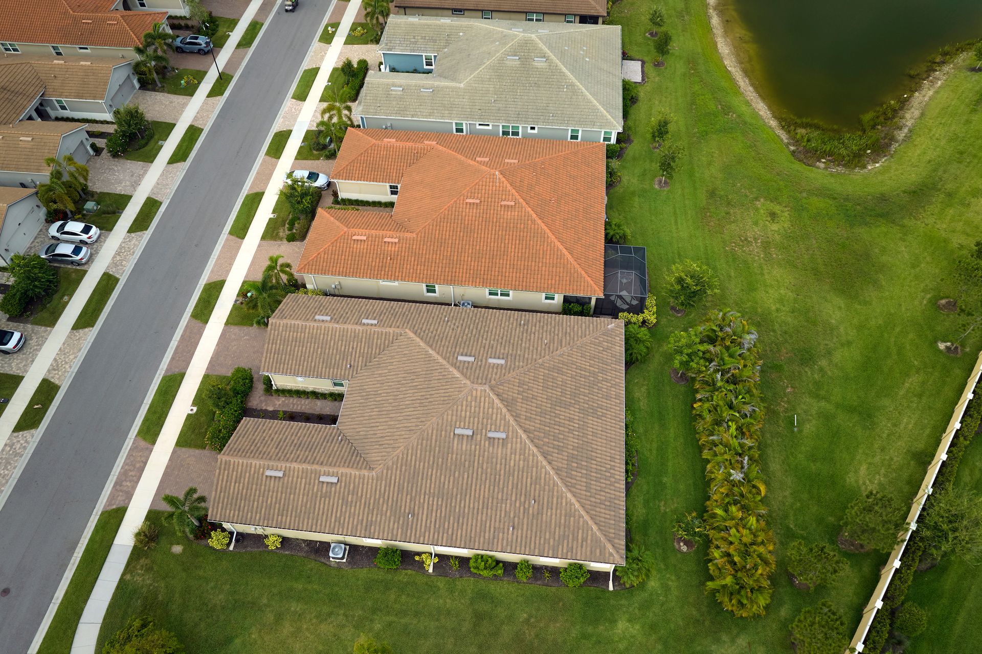 Aerial view of residential homes with different colored roofs along a street next to a grassy area and pond.