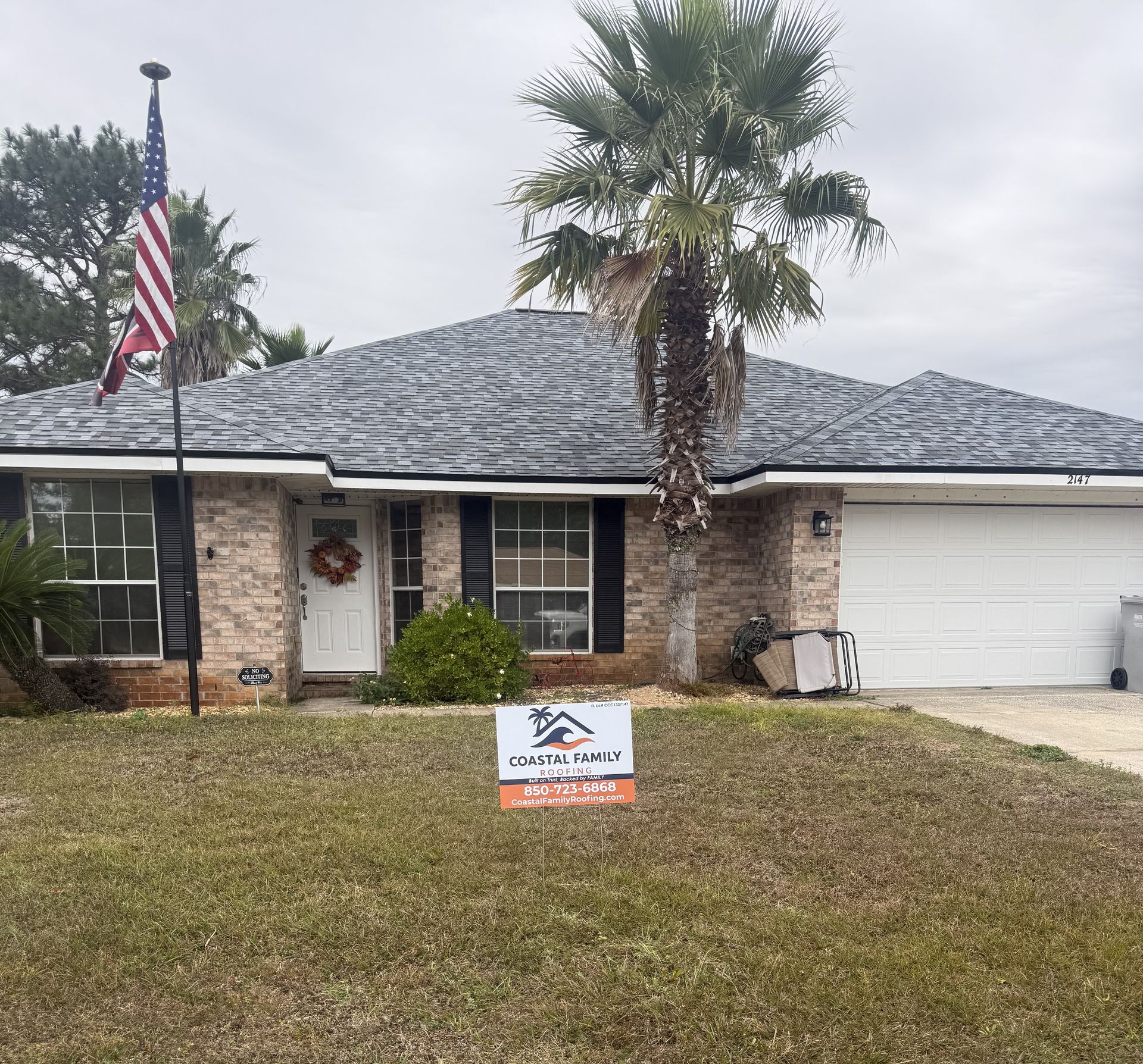 A single-story brick house with a new grey shingled roof, an American flag, and a yard sign, set under a cloudy sky.