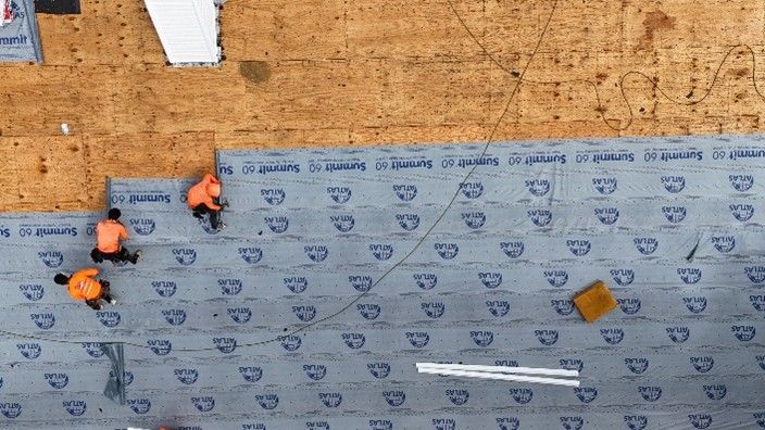 Three workers in orange gear install roofing underlayment on a wooden roof.