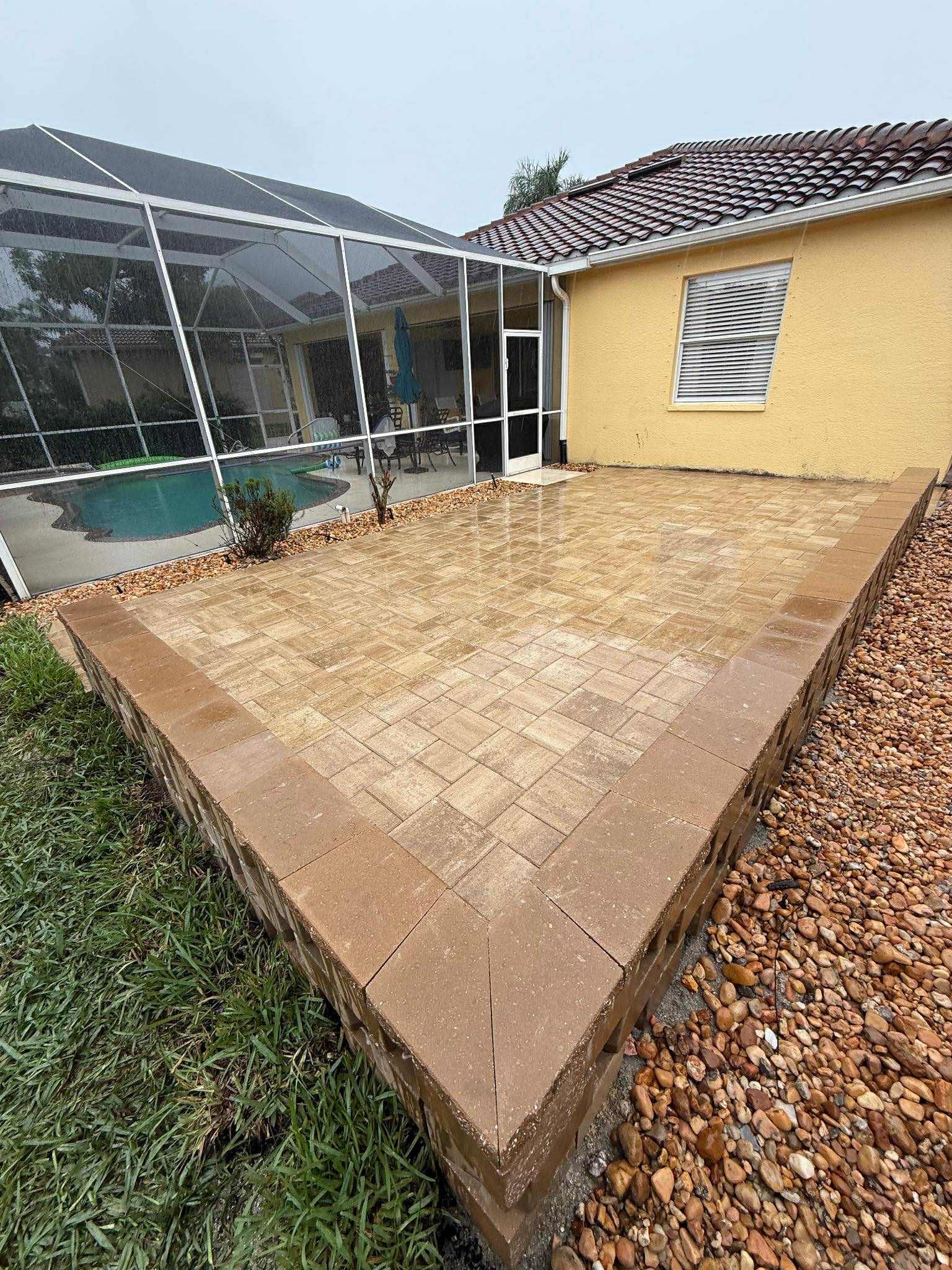 Patio with brick pavers, bordered by brown stones and retaining wall, next to a pool enclosure and house.