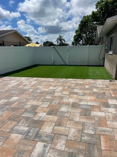 Patio with brick pavers, small patch of green turf, and white fence against a cloudy sky.