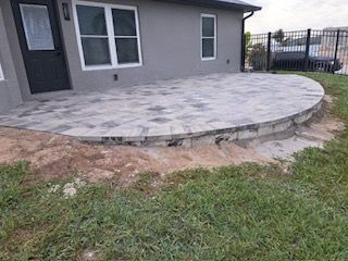 A paved patio, curved edge, built next to a gray house with windows and a door, surrounded by grass and dirt.