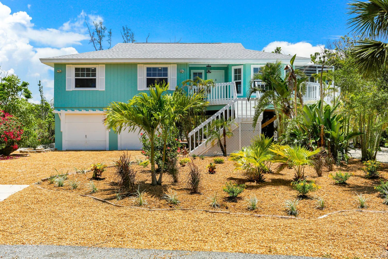 Coastal home with turquoise siding, white trim, and tropical landscaping under a blue sky.