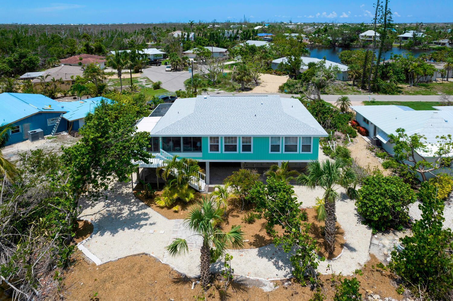 Aerial view of a teal-colored house with a gray roof in a tropical setting, surrounded by trees and other homes.