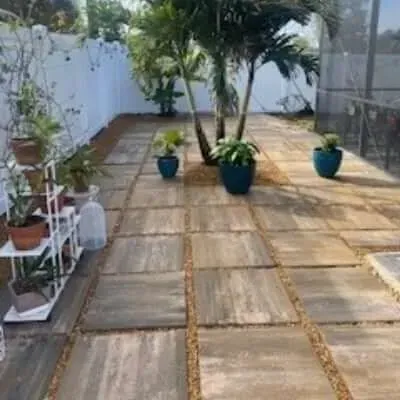 Patio with square pavers, gravel, plants, and a white fence in a sunny outdoor space.