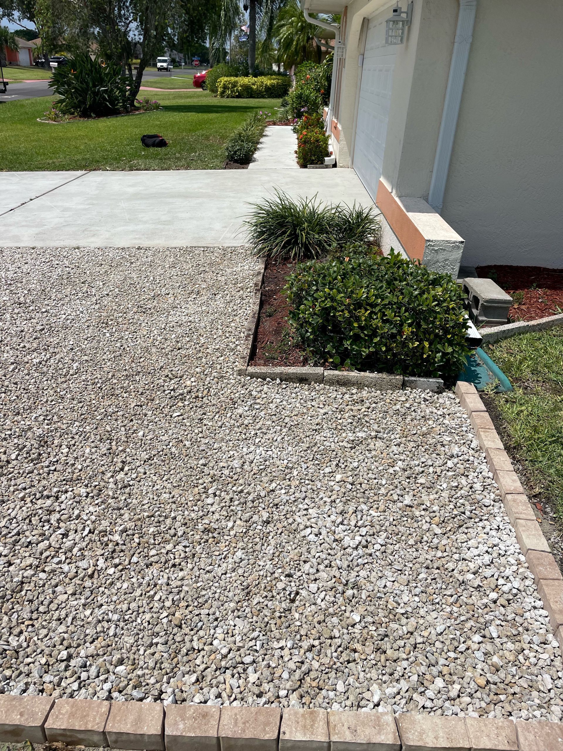 Gravel landscaping with a brick border, concrete walkway, and shrubs next to a light-colored house.